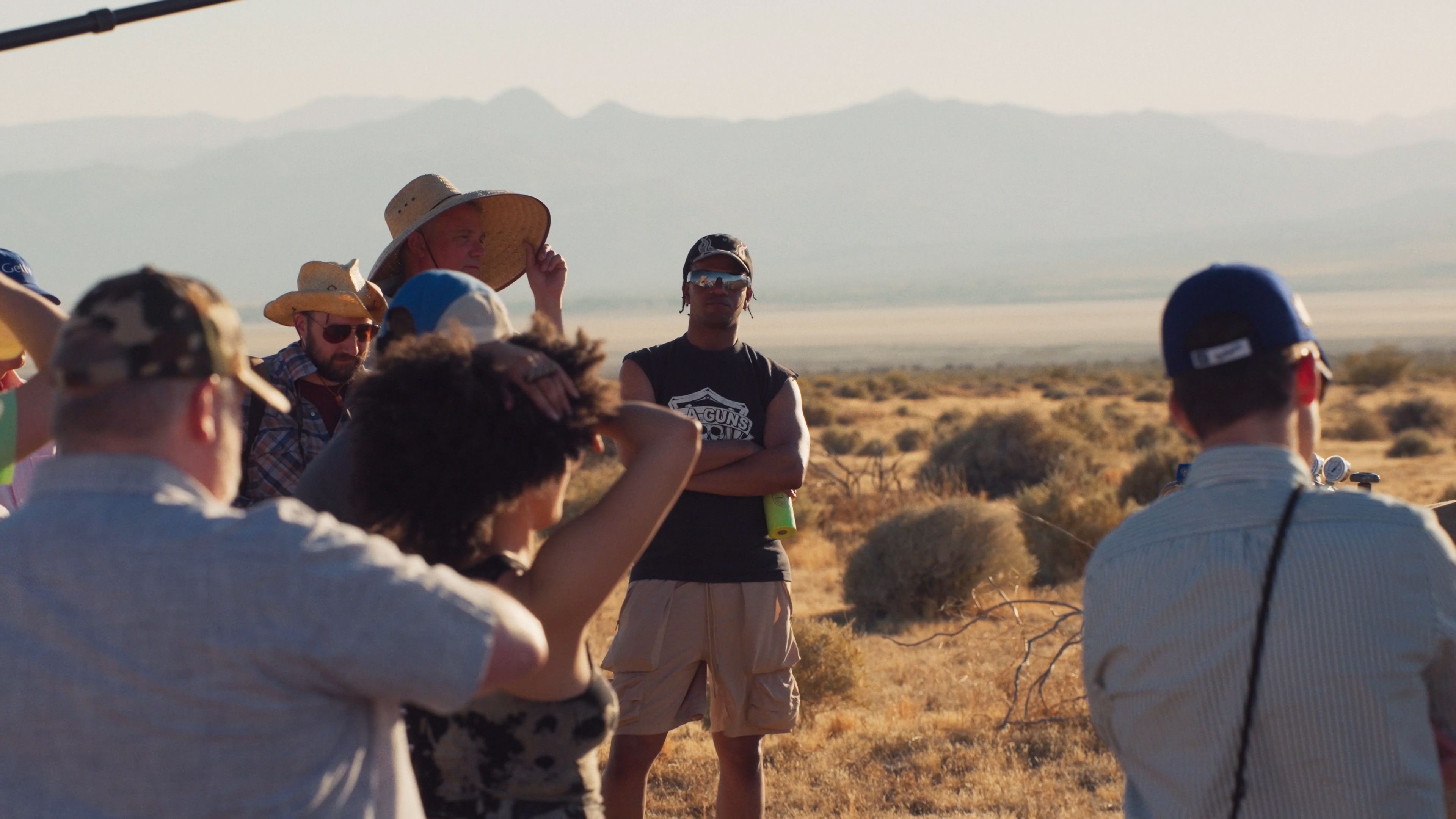 A group of people, some wearing hats, gather in a desert landscape with mountains in the background, engaged in a discussion or filming.