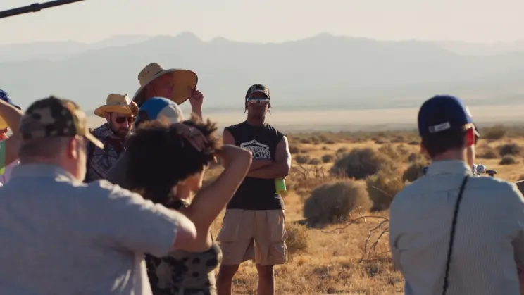 A group of people, some wearing hats, gather in a desert landscape with mountains in the background, engaged in a discussion or filming.