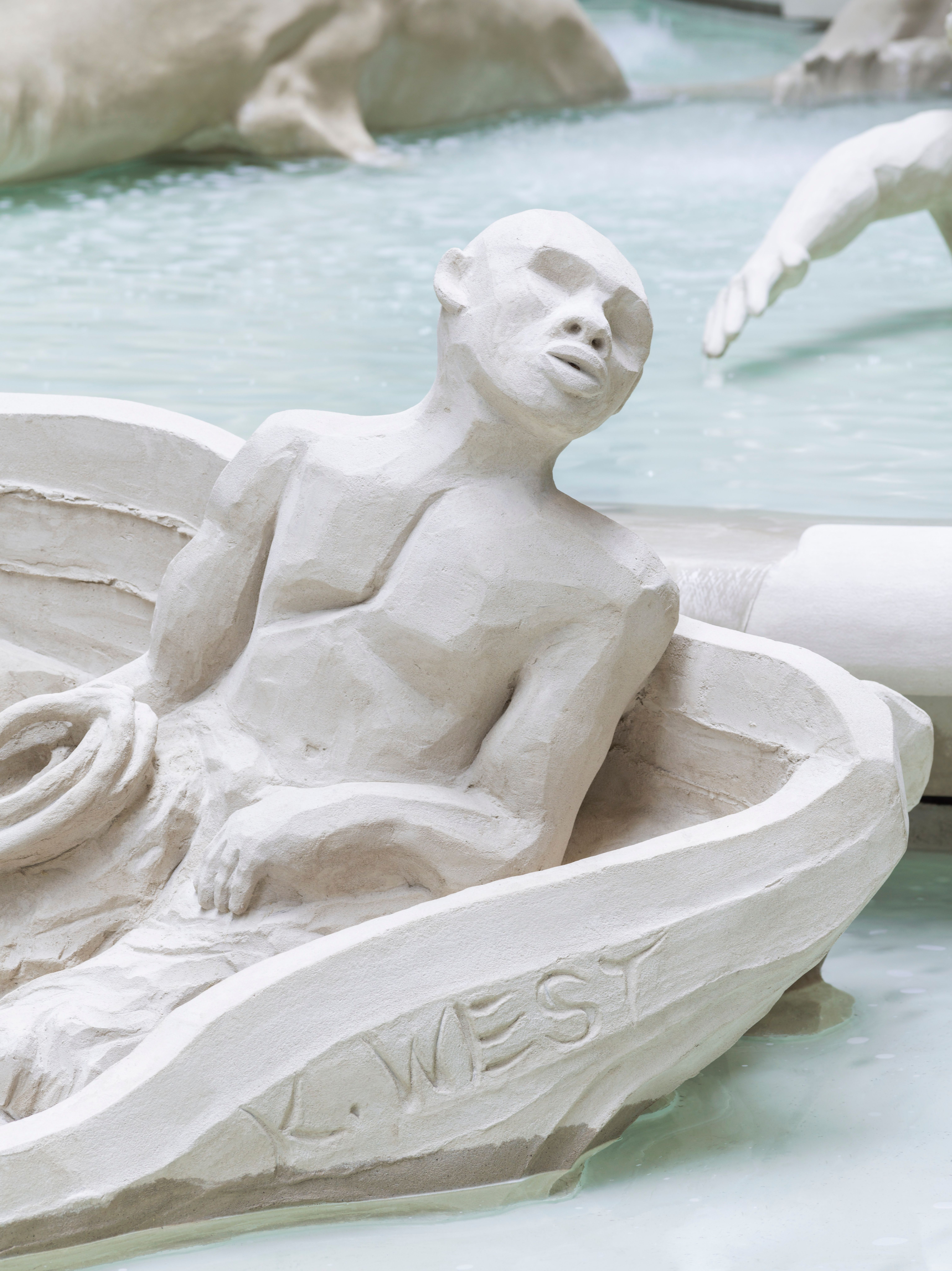 A segment of a detailed, white marble fountain depicts a figure seated in a boat. The setting is indoors with a neutral, s...
