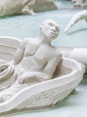 A segment of a detailed, white marble fountain depicts a figure seated in a boat. The setting is indoors with a neutral, s...