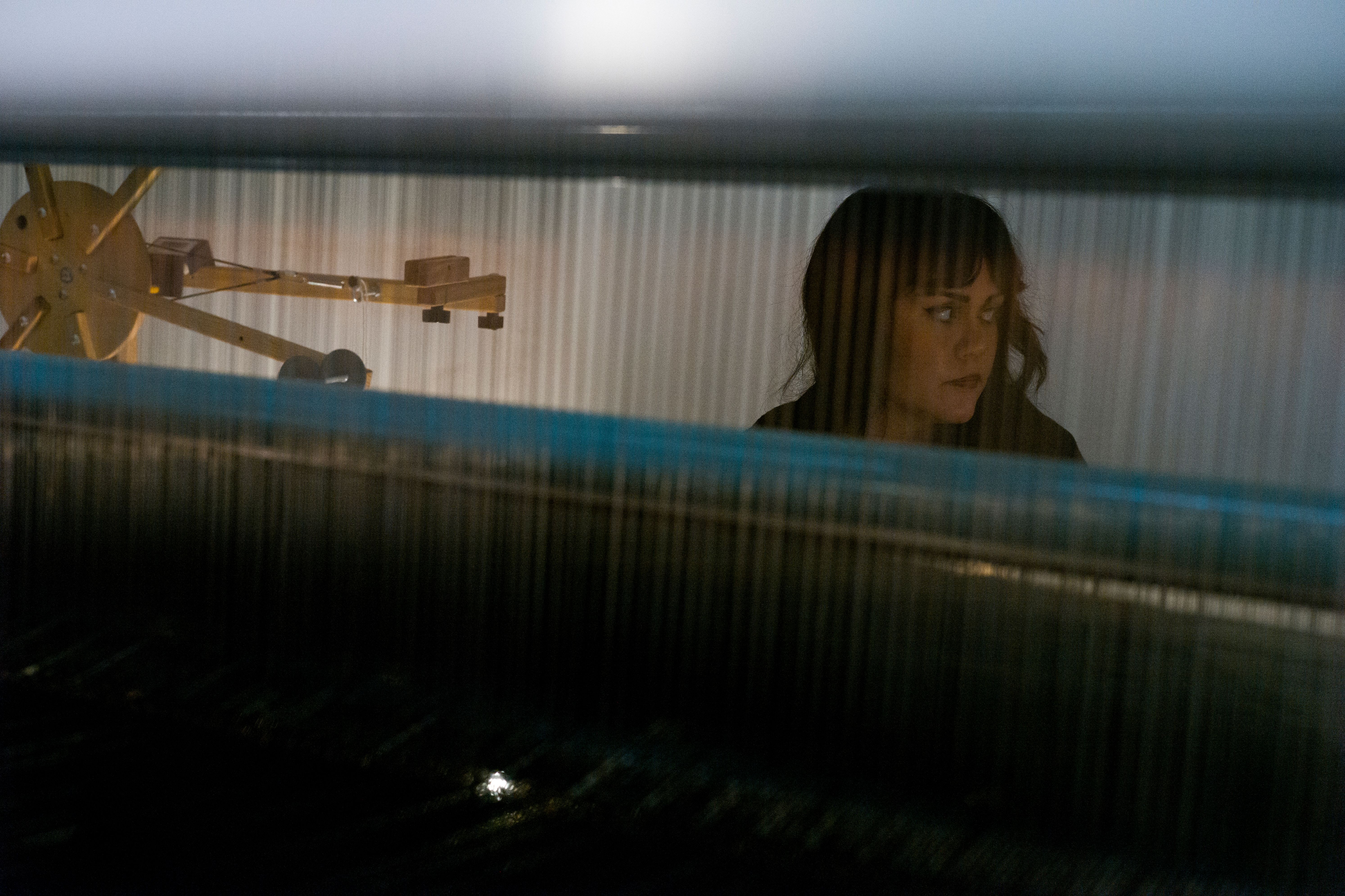A person viewed through the threads of a loom, focusing intently on their work.