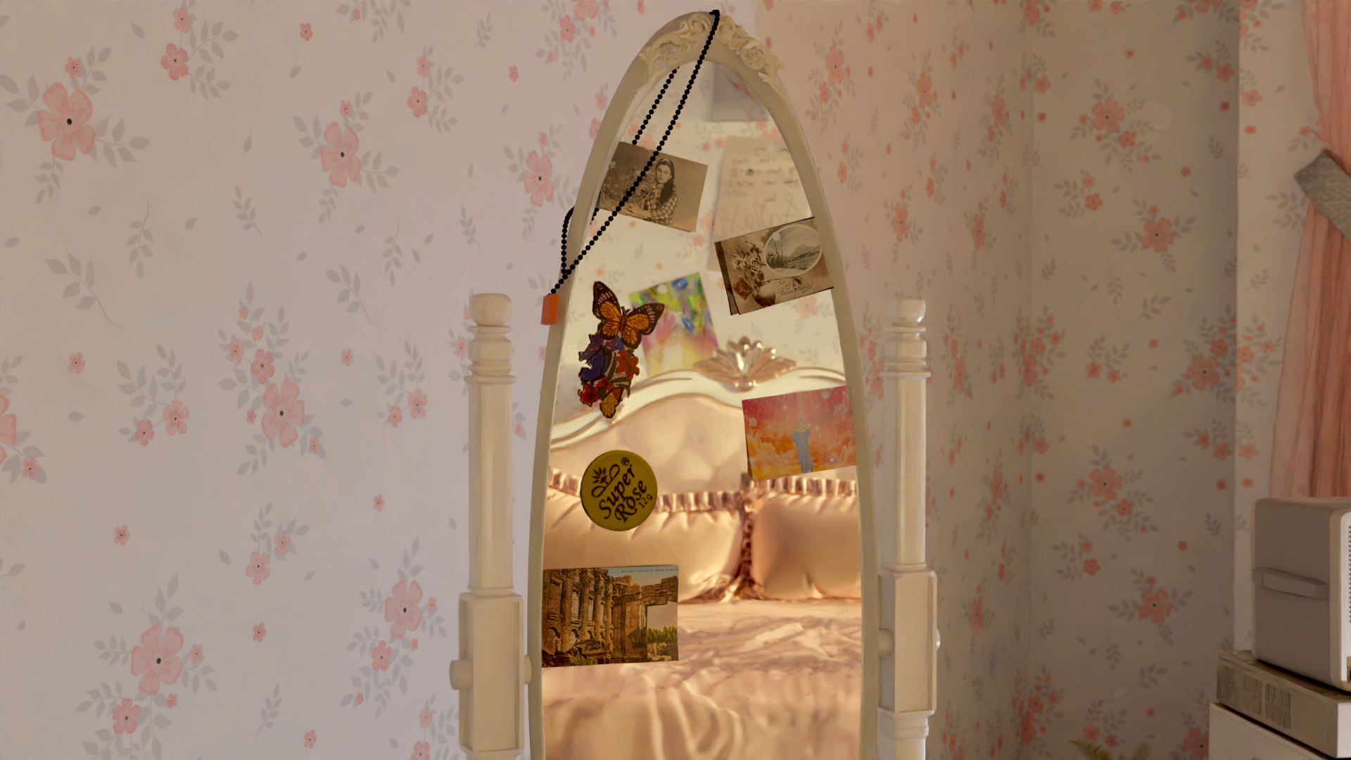 A mirror decorated with a hanging chain and photographs, with a glimpse of the bed and a floral wallpaper in the reflection.