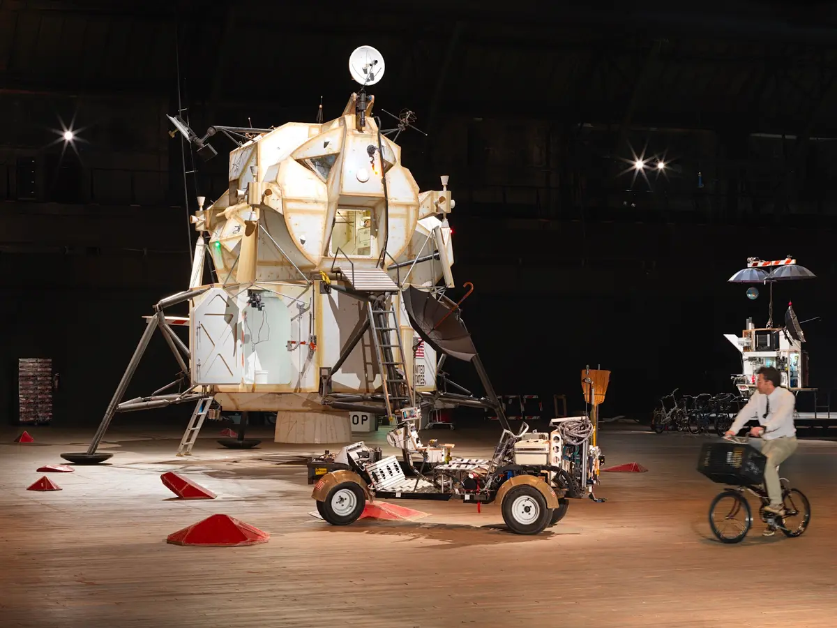 A man on a bicycle rides on a wooden floor, observing a display featuring a lunar lander and a lunar rover situated in a dimly lit, stage-like setting.