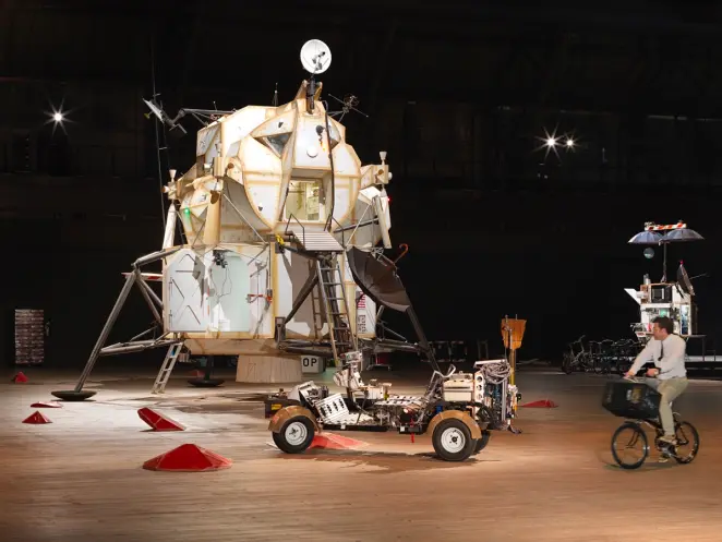 A man on a bicycle rides on a wooden floor, observing a display featuring a lunar lander and a lunar rover situated in a d...