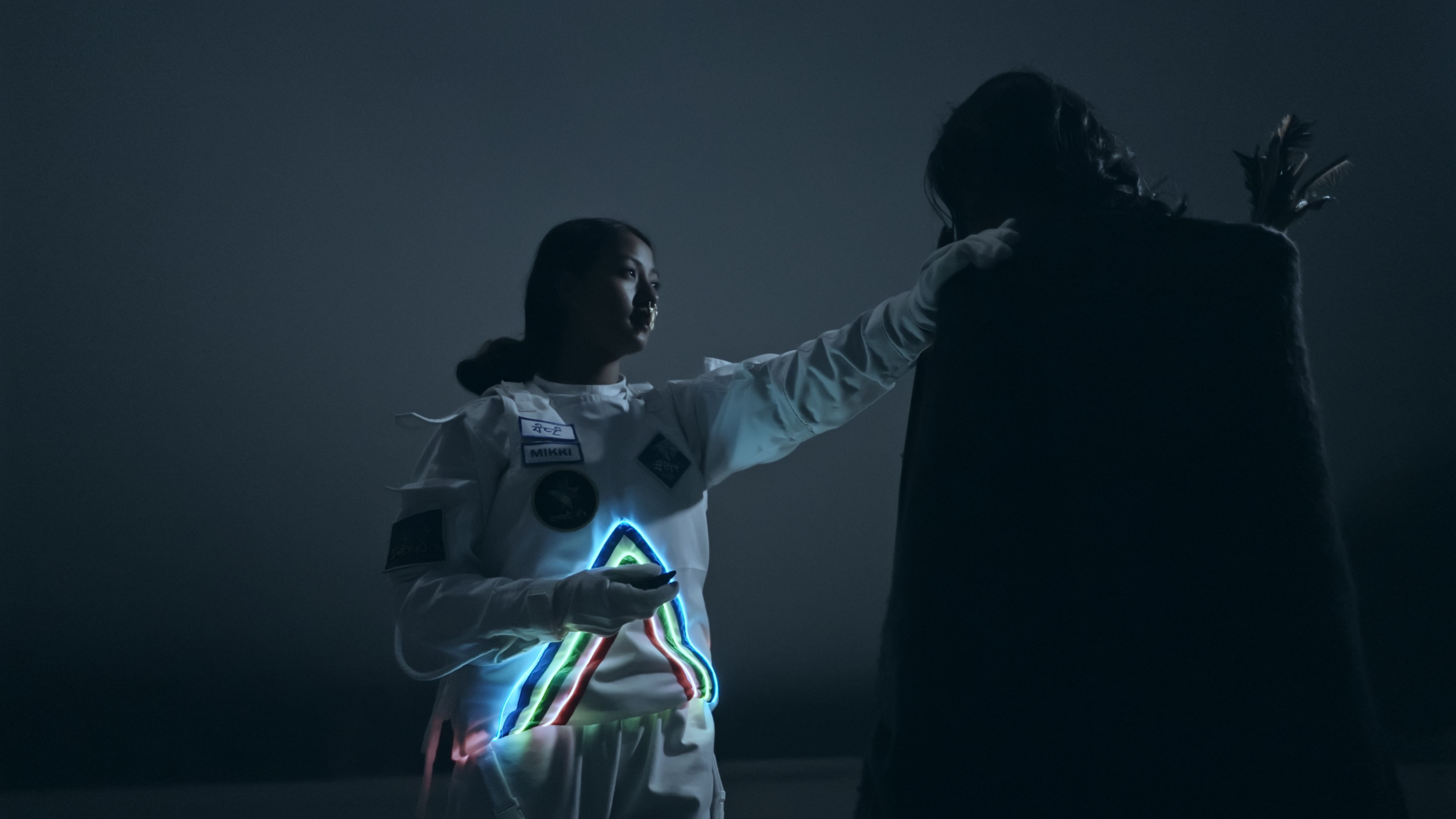 Two individuals stand facing each other at night. One is clad in a white suit decorated with neon lines forming a triangle.