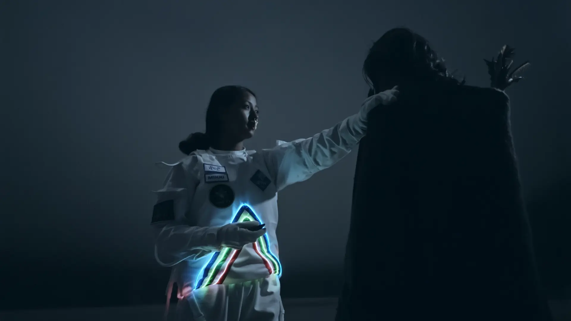 Two individuals stand facing each other at night. One is clad in a white suit decorated with neon lines forming a triangle.