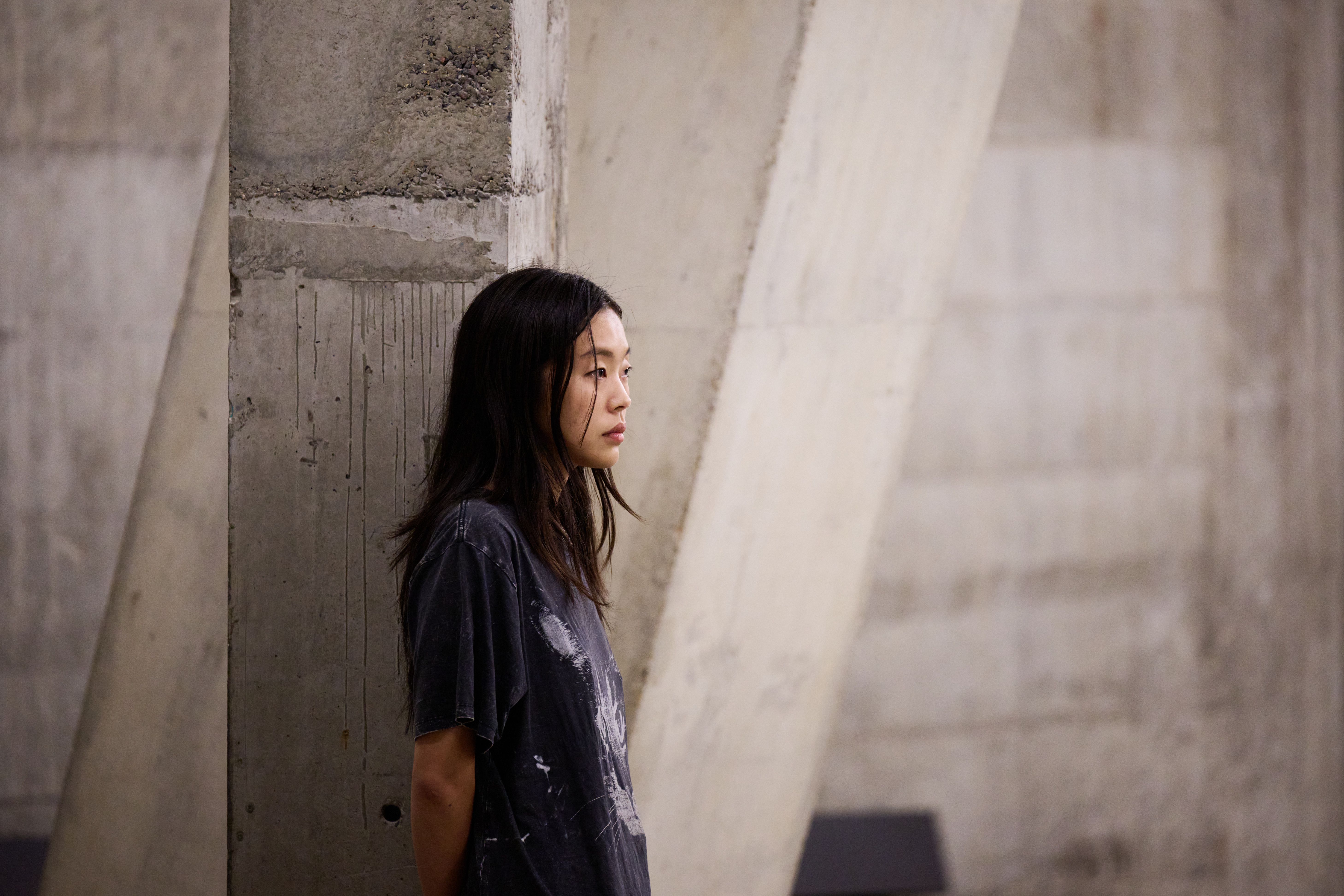 Artist stands against a concrete wall, looking contemplative, in a minimalist, industrial environment.