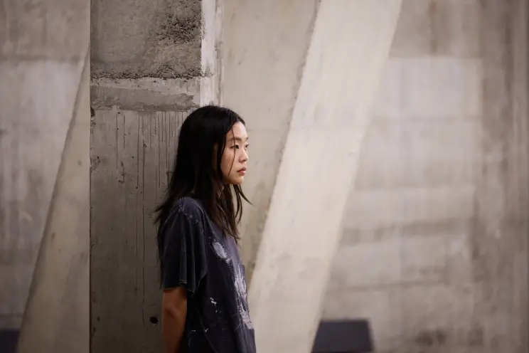 Artist stands against a concrete wall, looking contemplative, in a minimalist, industrial environment.