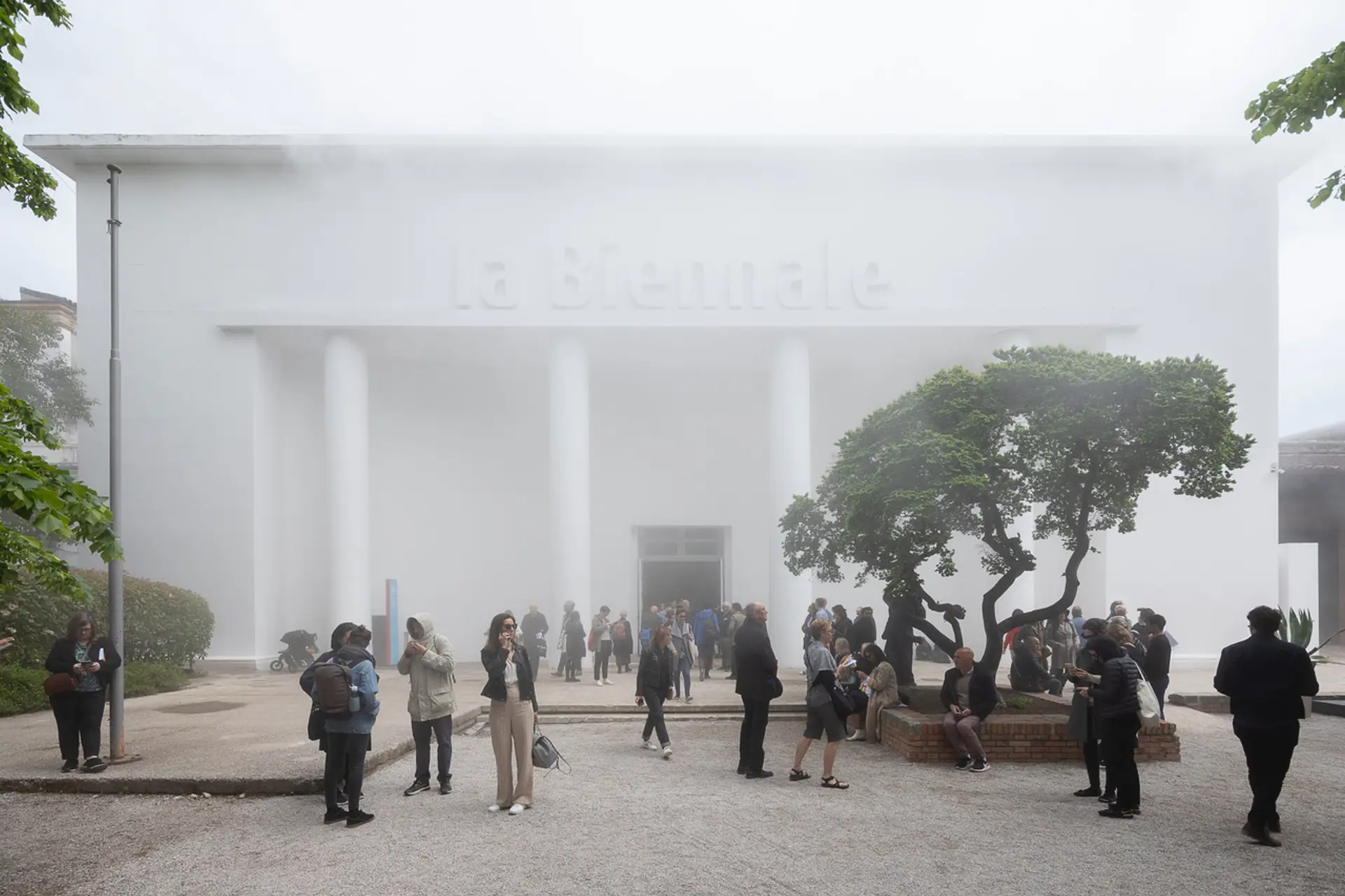 A bustling courtyard filled by a crowd of people in front of a grand white building, accentuated with architectural pillars.