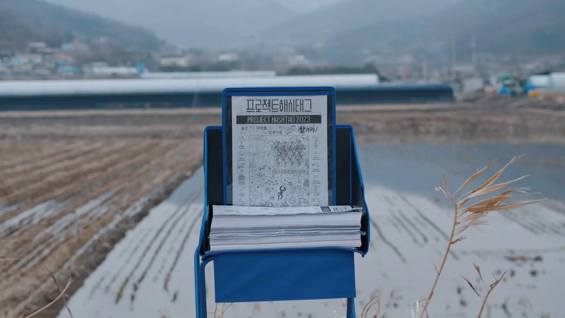 A newspaper titled "PROJECT HASHTAG 2023" in a blue stand with a blurred rural landscape and water in the background.
