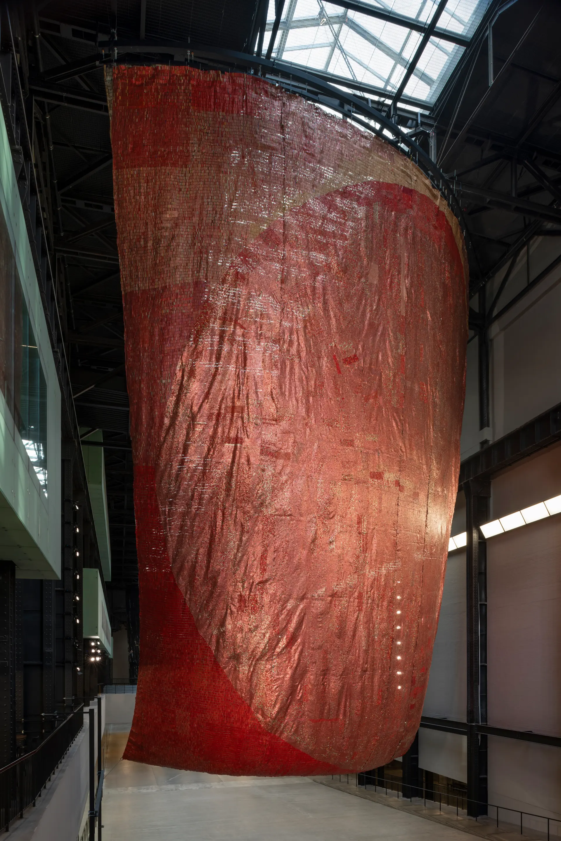 Large, draped red textile art installation hanging in a spacious industrial gallery, highlighted by natural light.