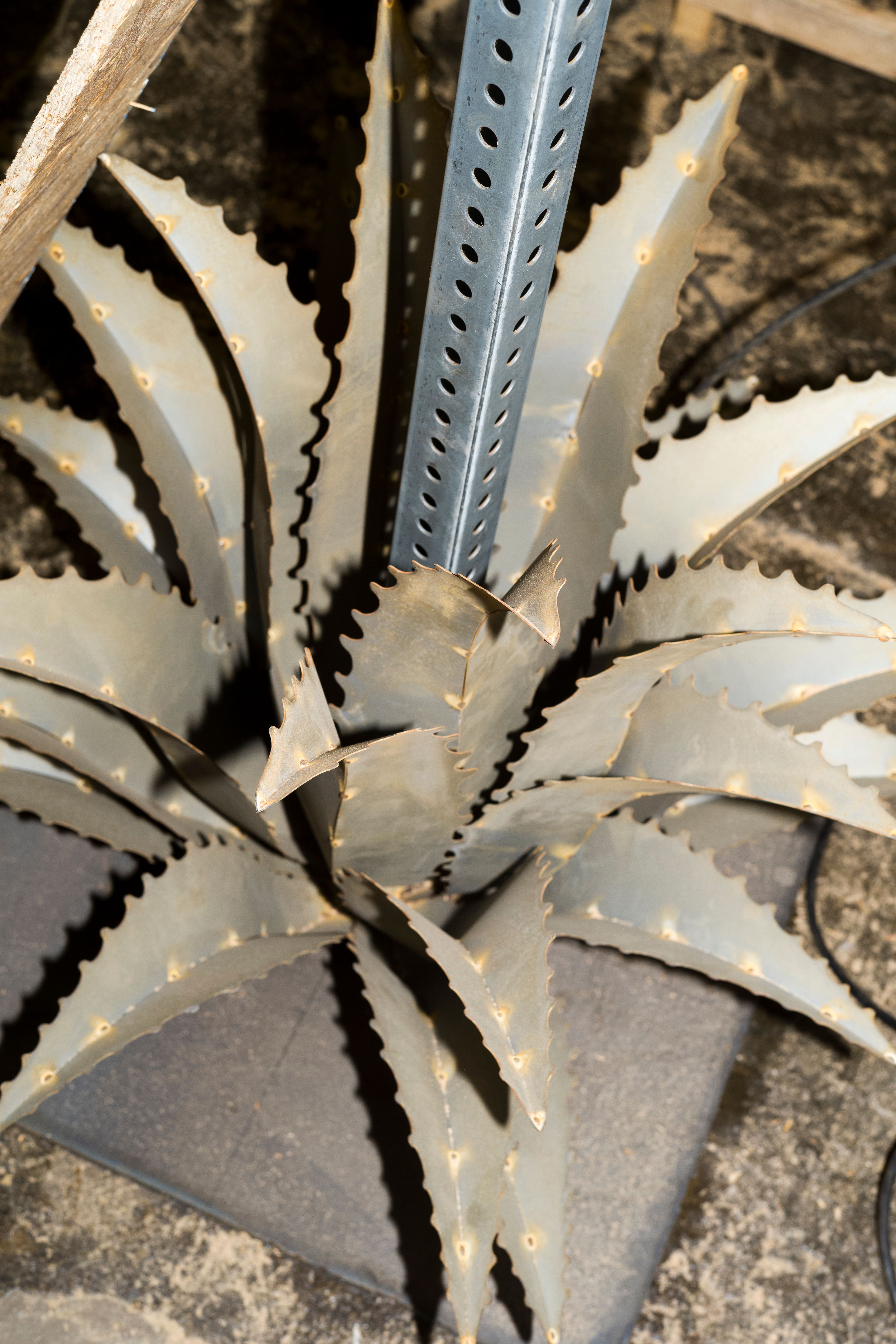 A metal sculpture resembling sharp agave leaves, positioned at the base of a perforated pole in an industrial setting.