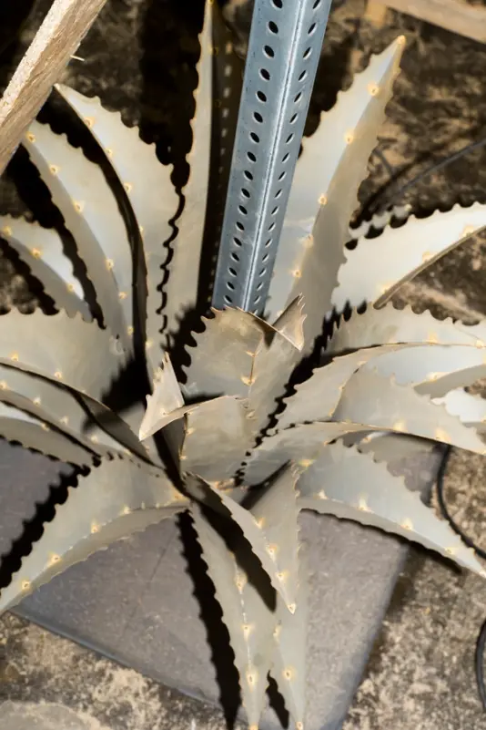 A metal sculpture resembling sharp agave leaves, positioned at the base of a perforated pole in an industrial setting.
