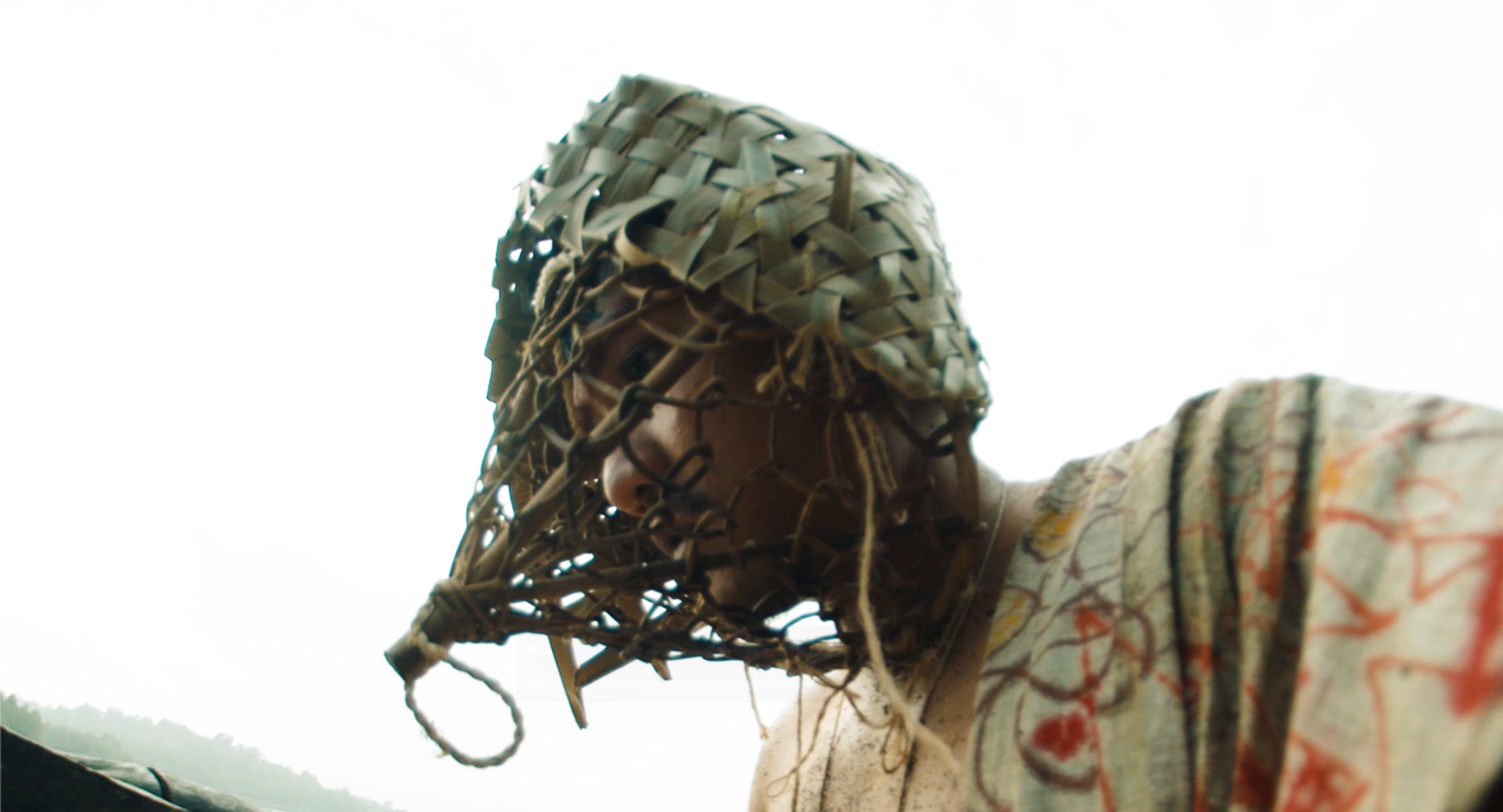 A person wearing a woven plant-fiber mask and painted robe looks down contemplatively outdoors.