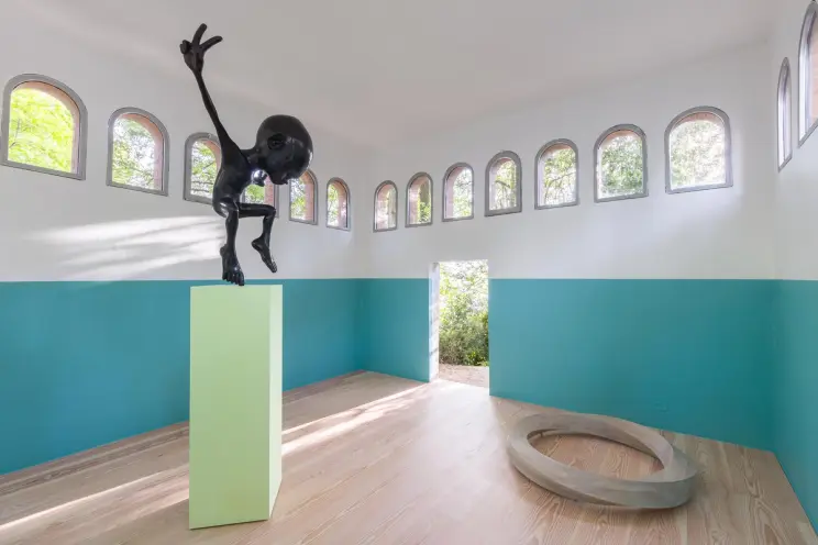 Art installation of a glossy black figure with outstretched limbs on a green pedestal in a room with teal and white walls,...