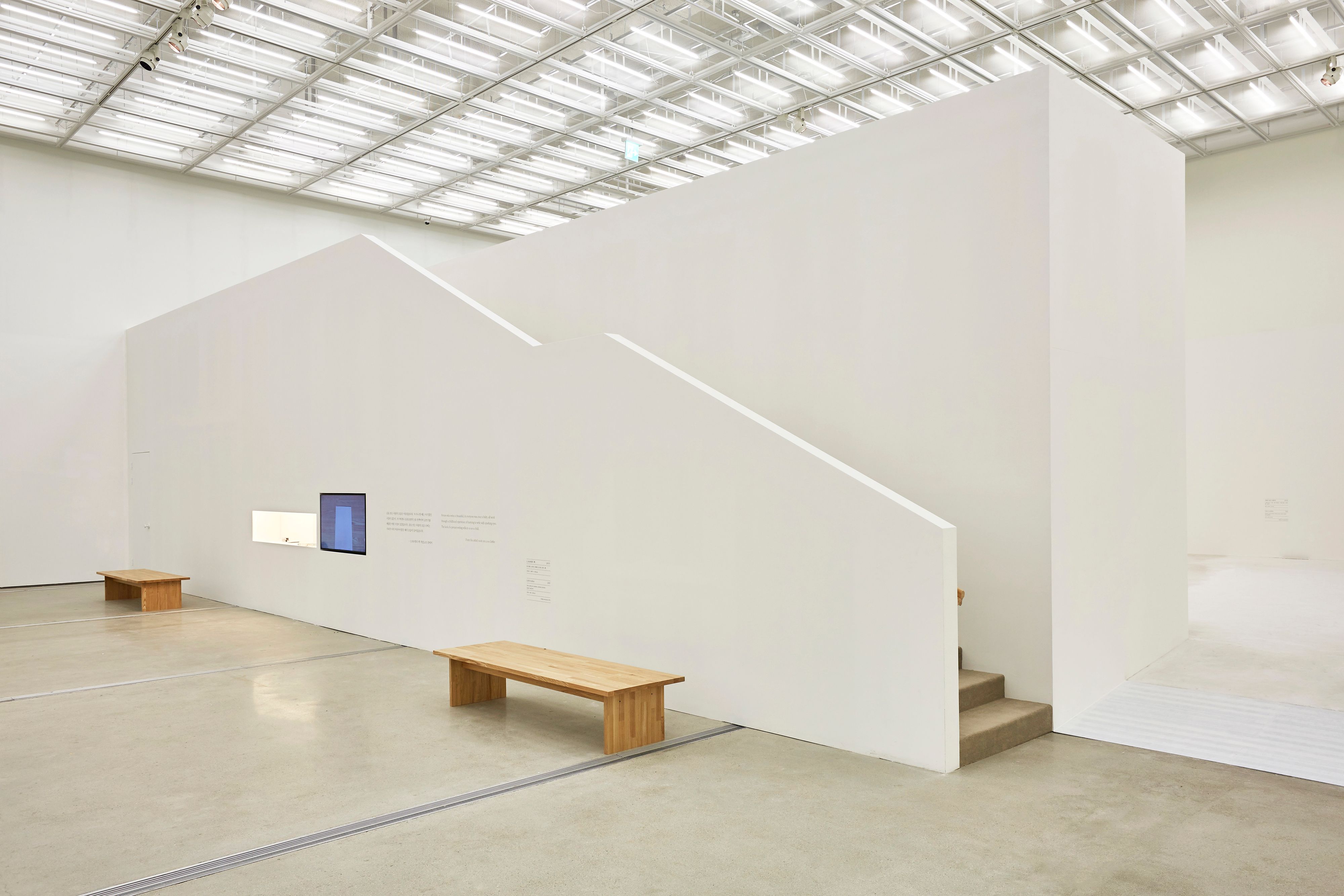 A gallery hall featuring two screens mounted on the wall, displaying content. Behind the screens, a staircase ascends. In ...