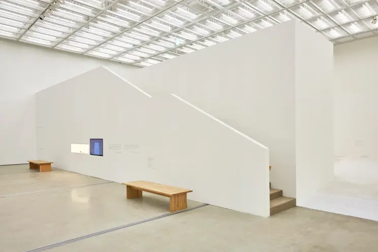 A gallery hall featuring two screens mounted on the wall, displaying content. Behind the screens, a staircase ascends. In ...