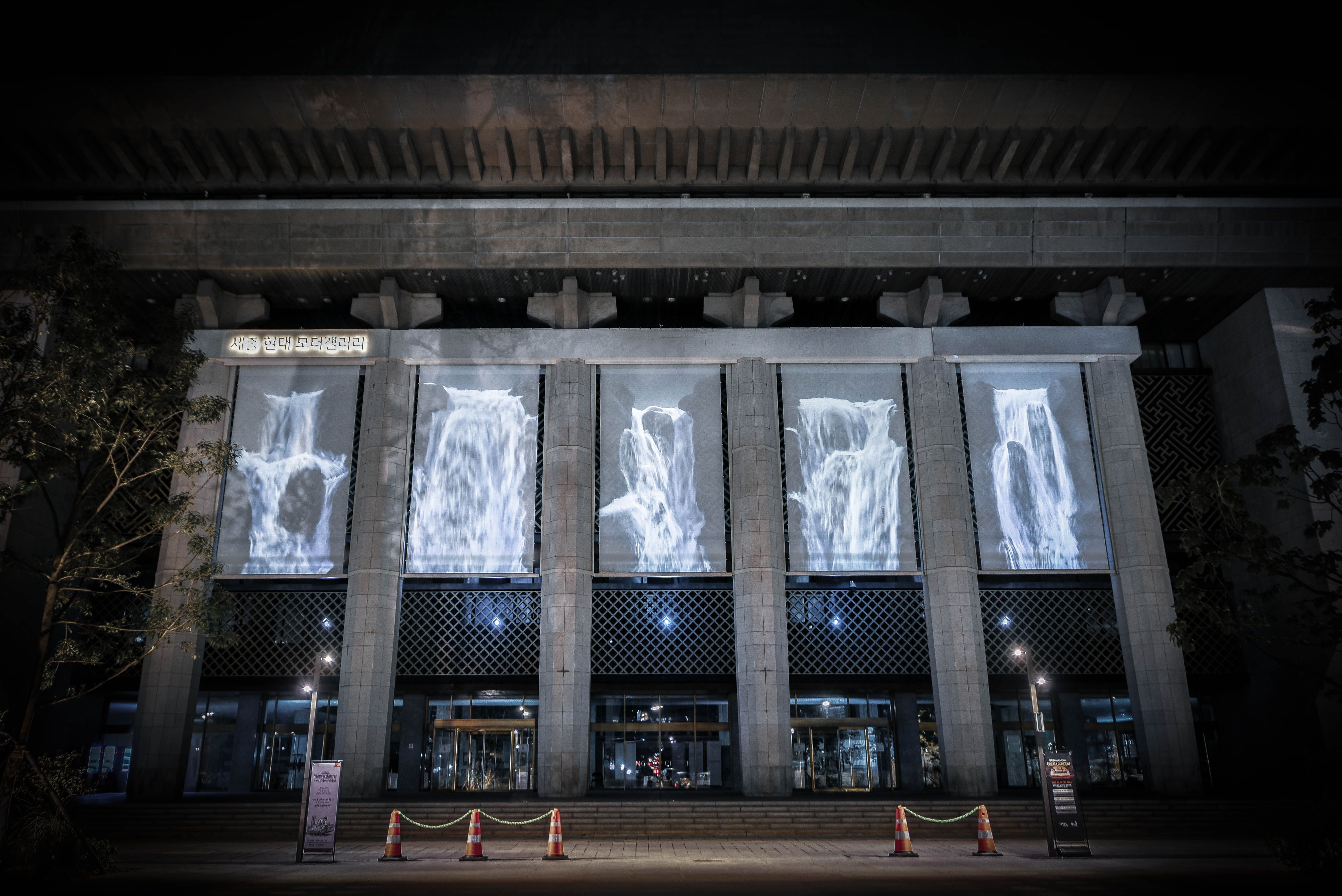The façade of a large building illuminated with projected patterned greys. A line of traffic cones in front of the buildin...