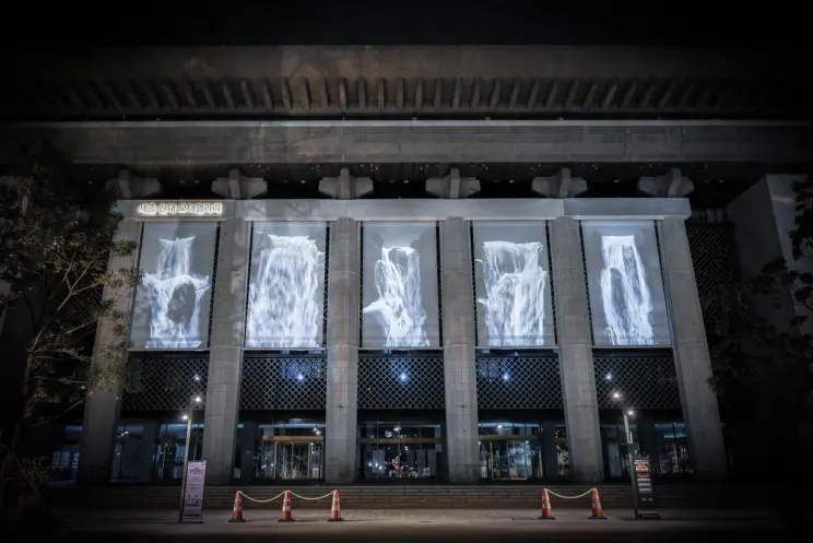 The façade of a large building illuminated with projected patterned greys. A line of traffic cones in front of the buildin...