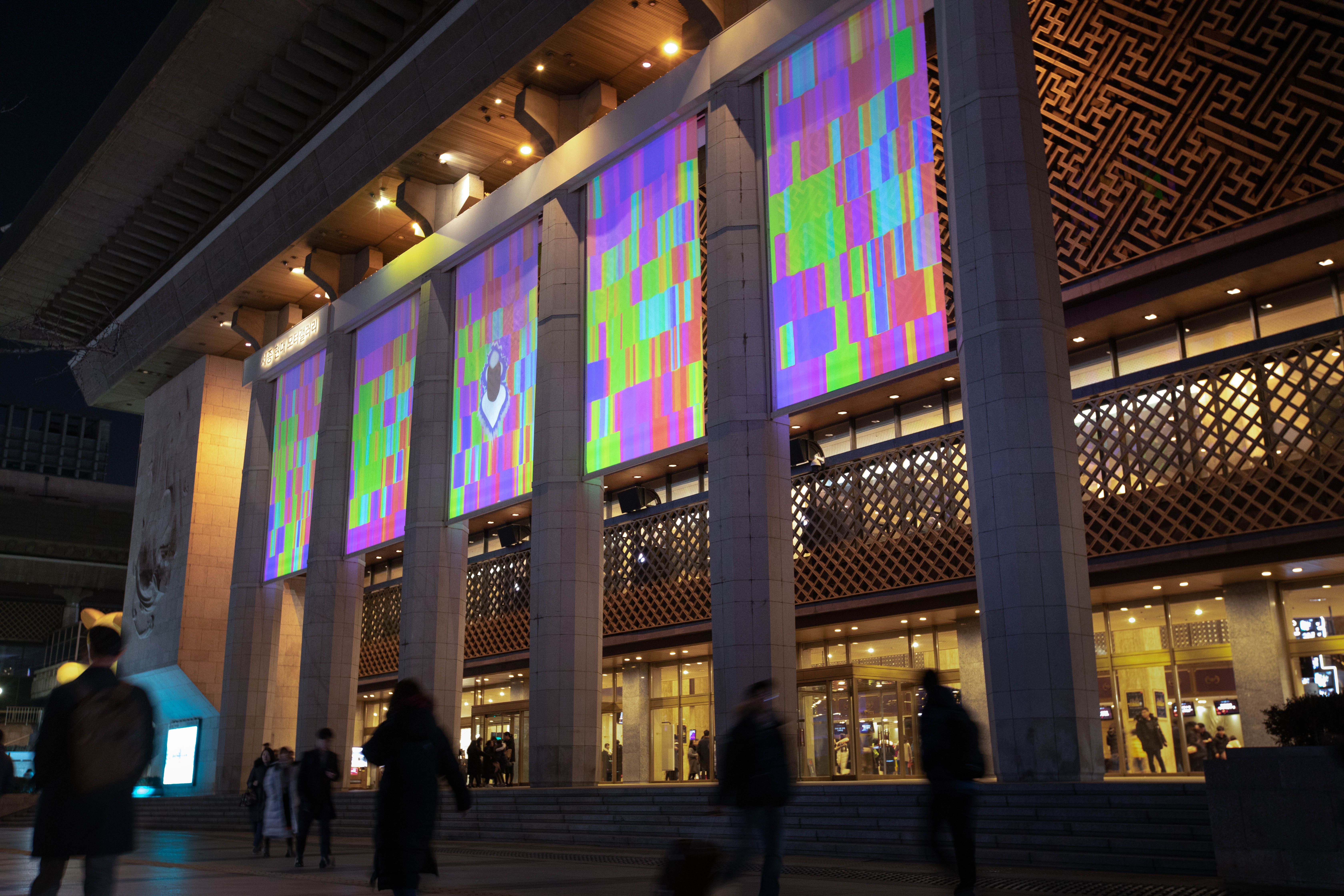 Building facade with colorful digital art projections, people walking by at night.