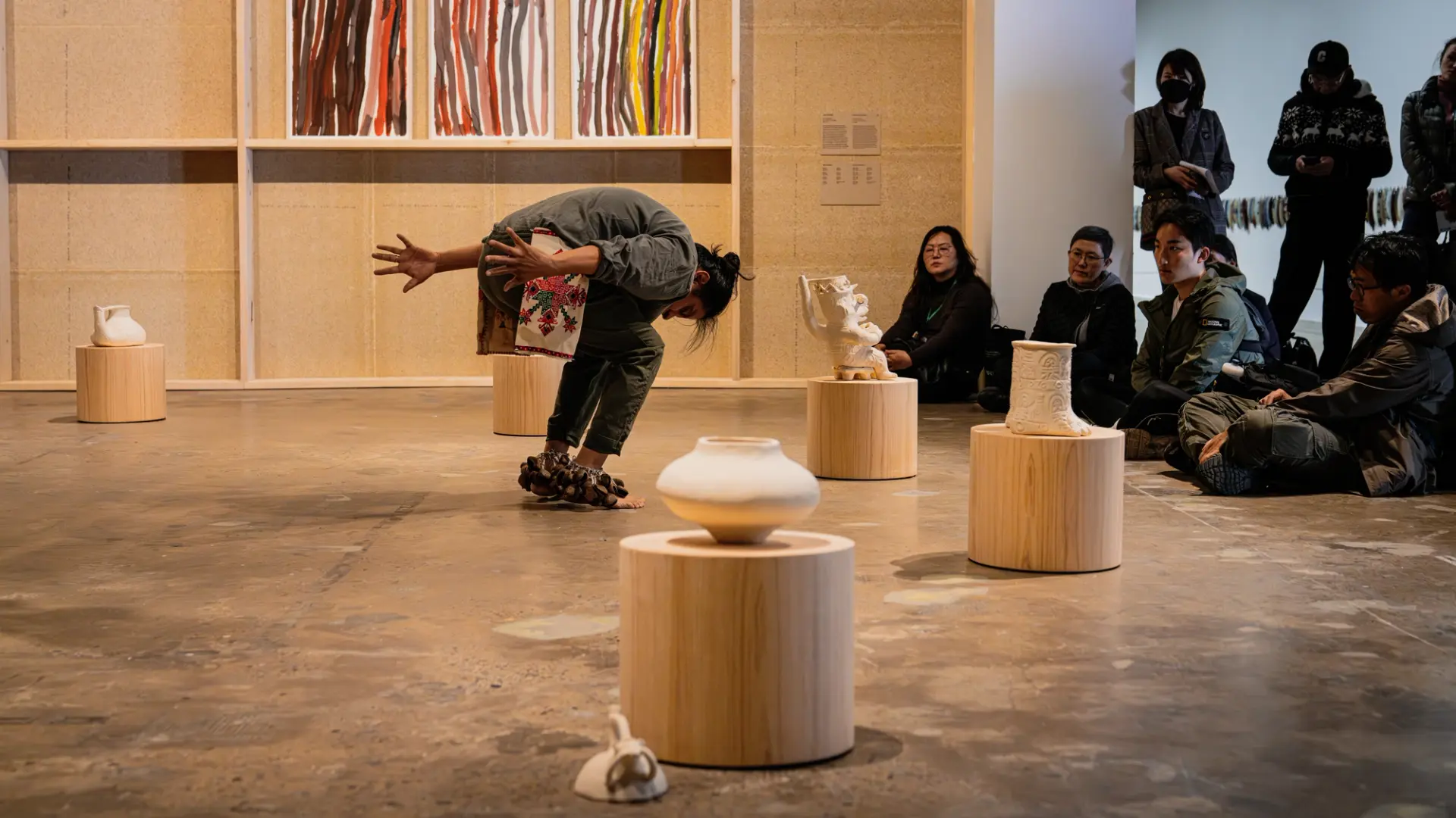 Person performs amid various art pieces, while an audience seated on the floor watches attentively.