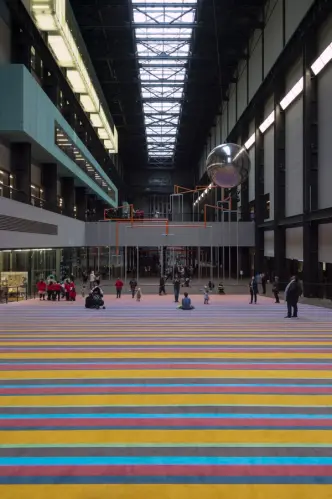 A vibrant gallery space filled with horizontal multicolored lines on the floor. A spherical object is suspended from the c...