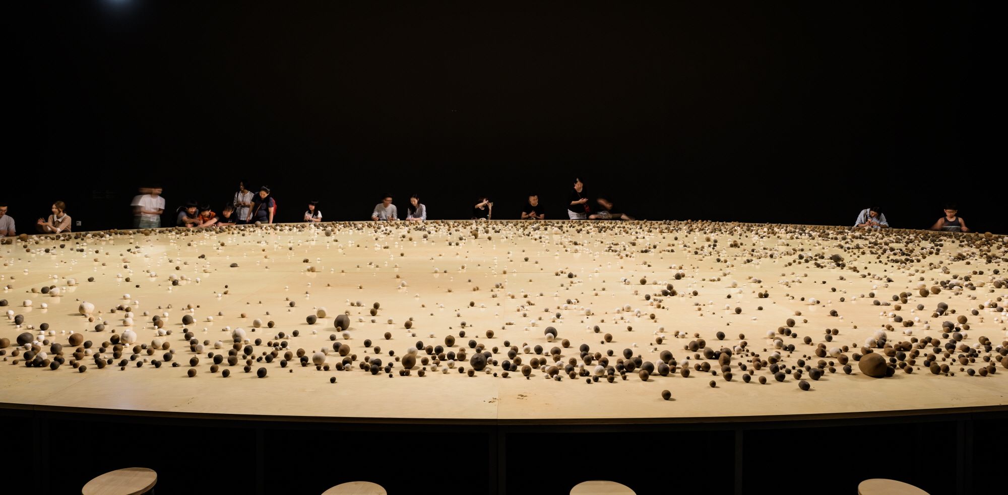 An enormous round wooden table populated with various sized round rocks, under a dimmed lighting. Several individuals stan...