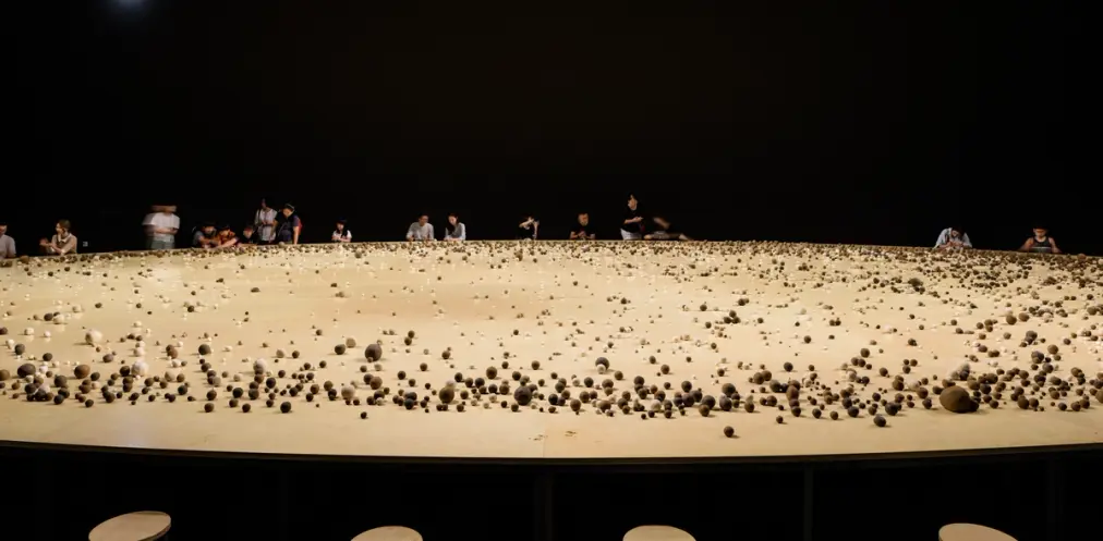 An enormous round wooden table populated with various sized round rocks, under a dimmed lighting. Several individuals stan...