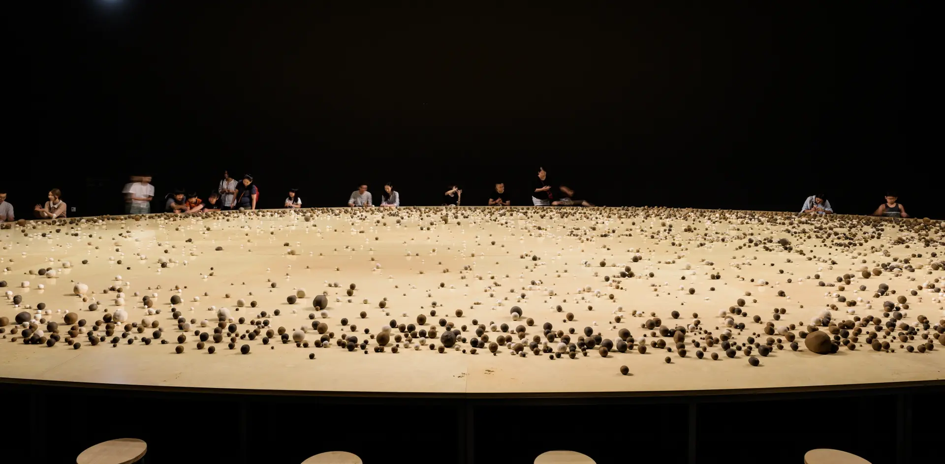 An enormous round wooden table populated with various sized round rocks, under a dimmed lighting. Several individuals stan...