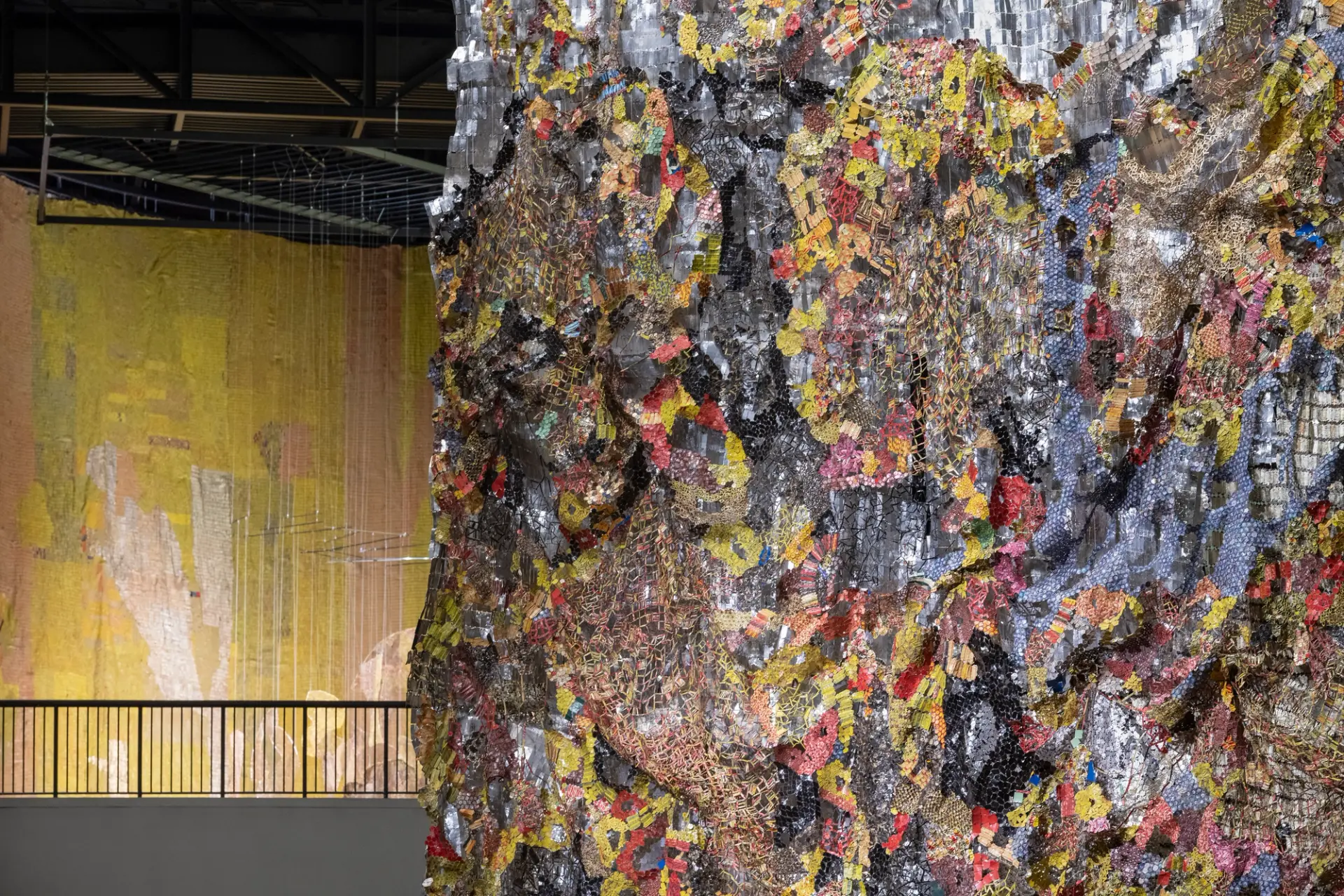 Textured and colorful collage art installation displayed in a gallery, juxtaposed against a large yellow hanging backdrop,...