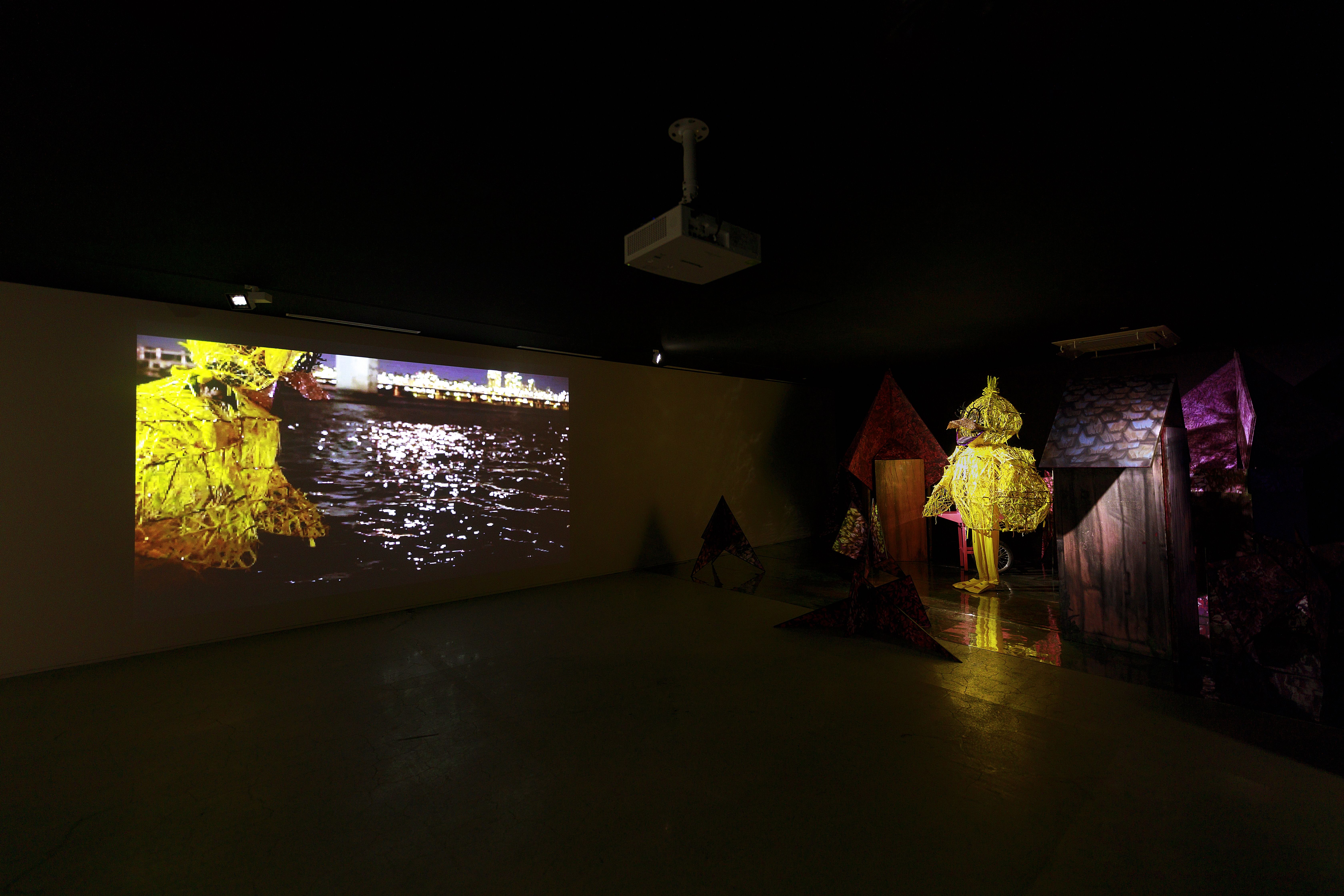 Within a dimly lit gallery, a large bird-like sculpture occupies the room, accompanied by projected images on the adjacent...