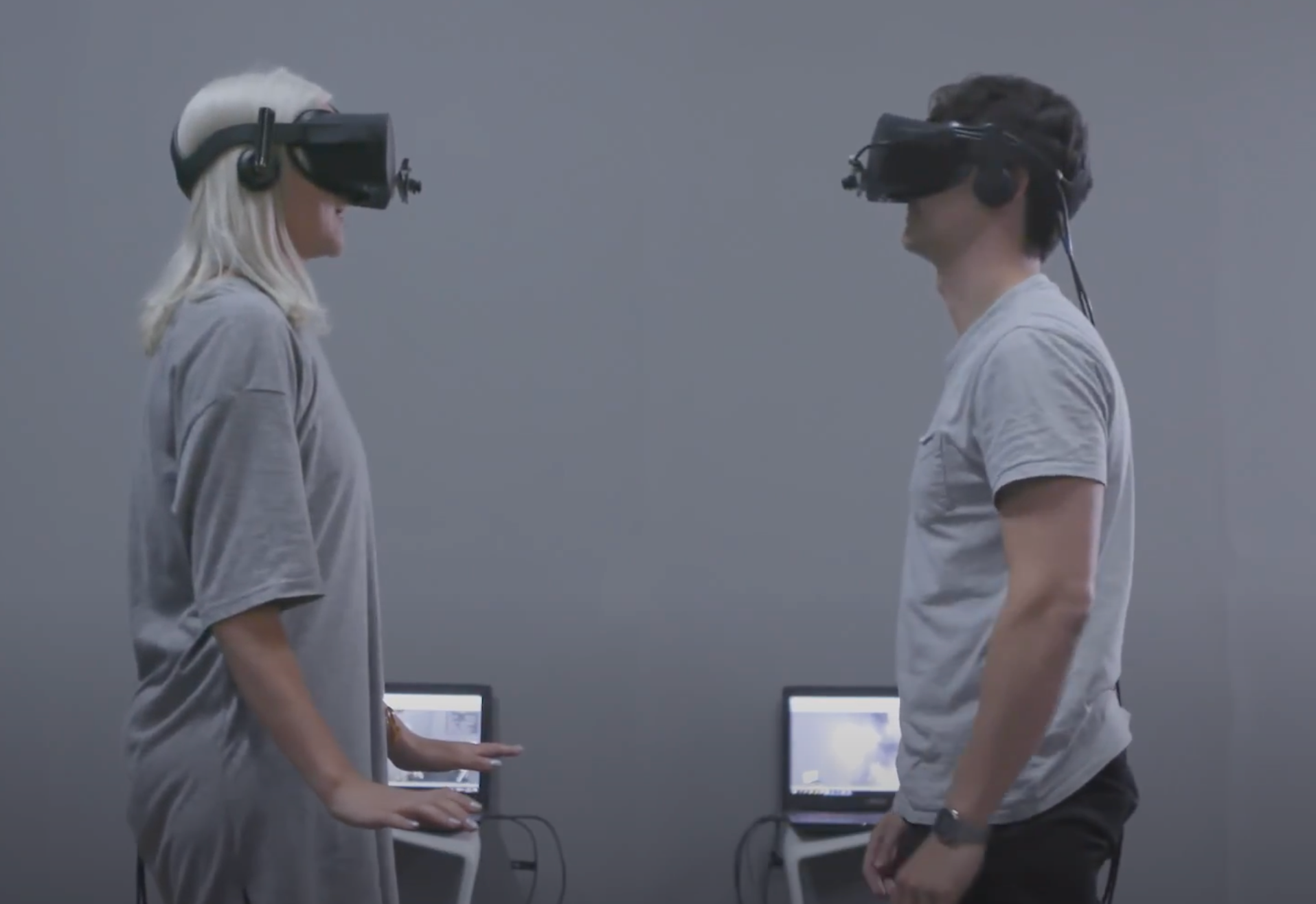 Two individuals wearing grey shirts stand facing each other, each immersed in their own virtual reality headsets. Behind t...
