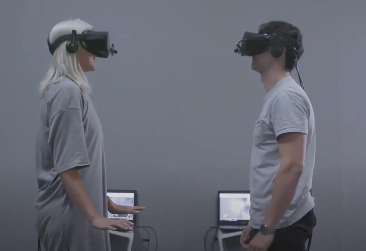 Two individuals wearing grey shirts stand facing each other, each immersed in their own virtual reality headsets. Behind them, two laptops rest on a solid grey wall.