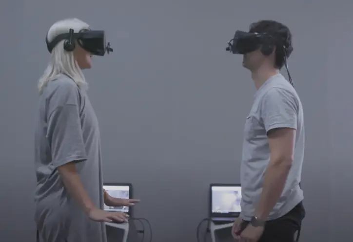 Two individuals wearing grey shirts stand facing each other, each immersed in their own virtual reality headsets. Behind t...