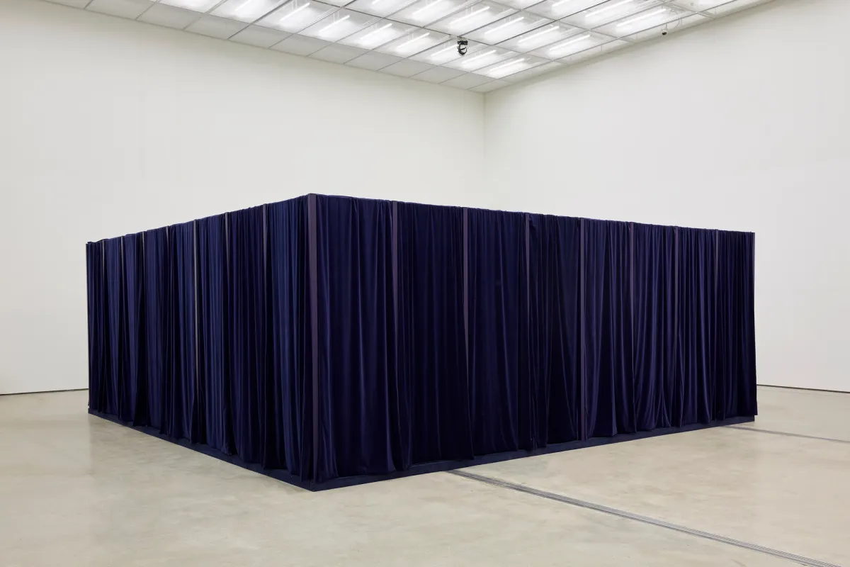 A sizable cube draped in dark blue fabric dominates the center of a brightly lit exhibition room.