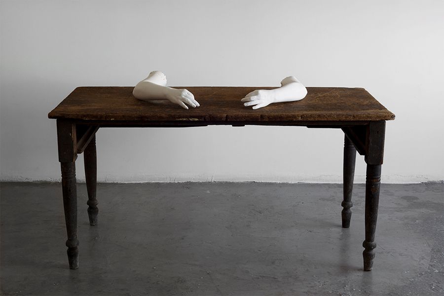 An antiquated wooden desk sits on a grey concrete floor with a white sculpted artwork positioned as if they are writing on...