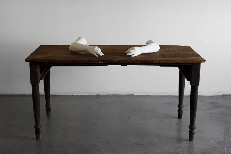 An antiquated wooden desk sits on a grey concrete floor with a white sculpted artwork positioned as if they are writing on...