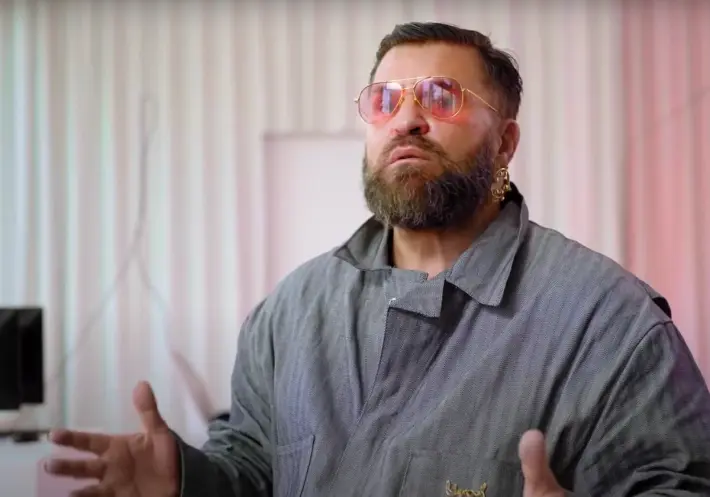 A person in a grey shirt and red glasses gesturing while speaking to the camera in a well-lit room.