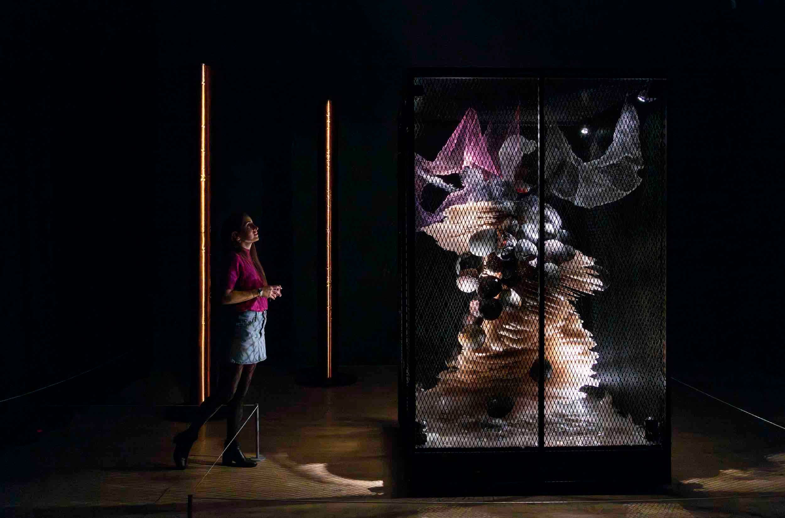 "A woman in a magenta top and light blue skirt stands in a dark gallery space, gazing at a large sculptural installation enclosed in a black metal mesh cage. The sculpture is an abstract, organic form composed of layered, flowing fabrics and various spherical and reflective objects. Warm vertical light bars flank the installation, creating a dramatic and immersive atmosphere."