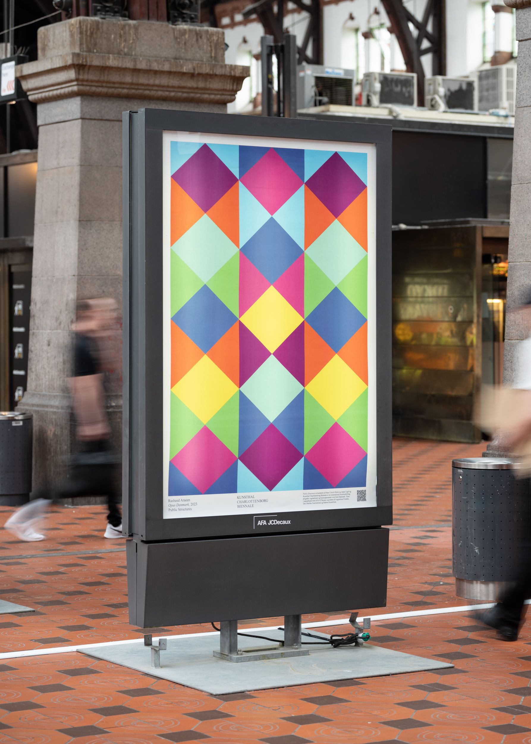 A small vertical billboard showing an exhibition poster showing a colorful checkered art work.