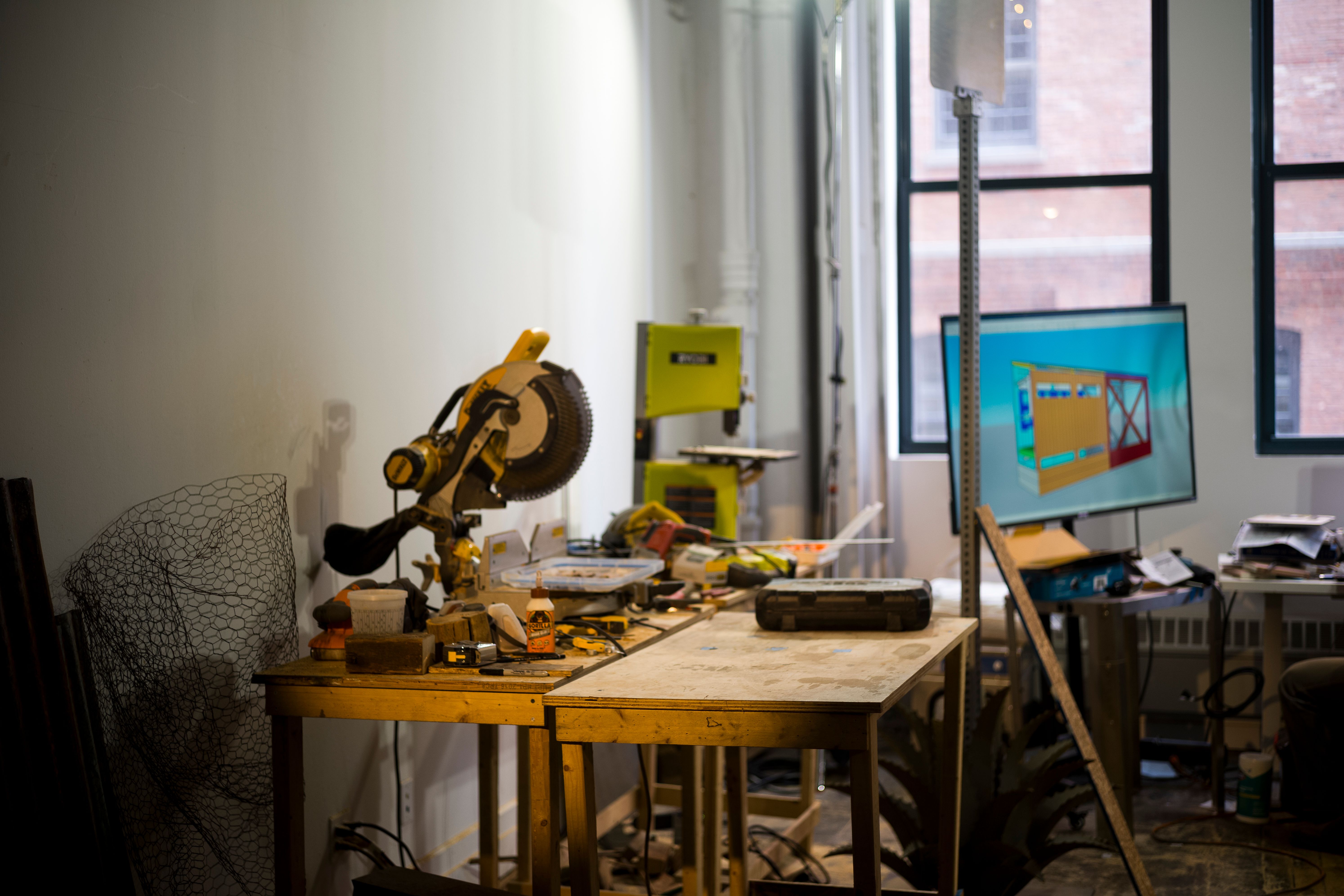 A cluttered workshop table with tools and equipment, including a saw and various materials, in a well-lit room.