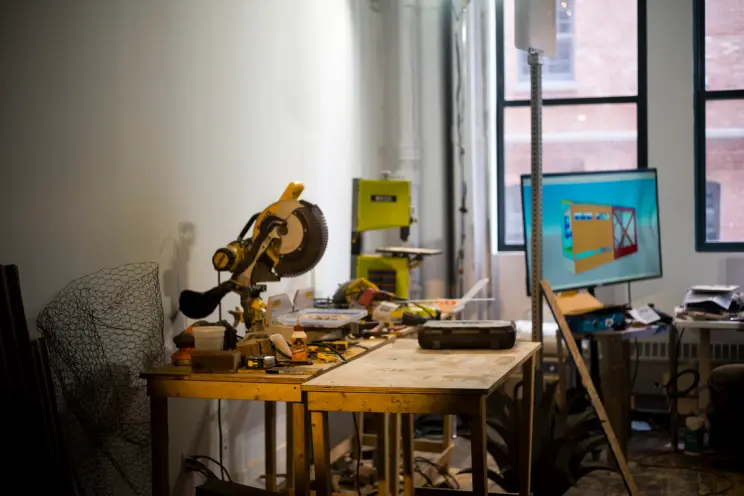 A cluttered workshop table with tools and equipment, including a saw and various materials, in a well-lit room.