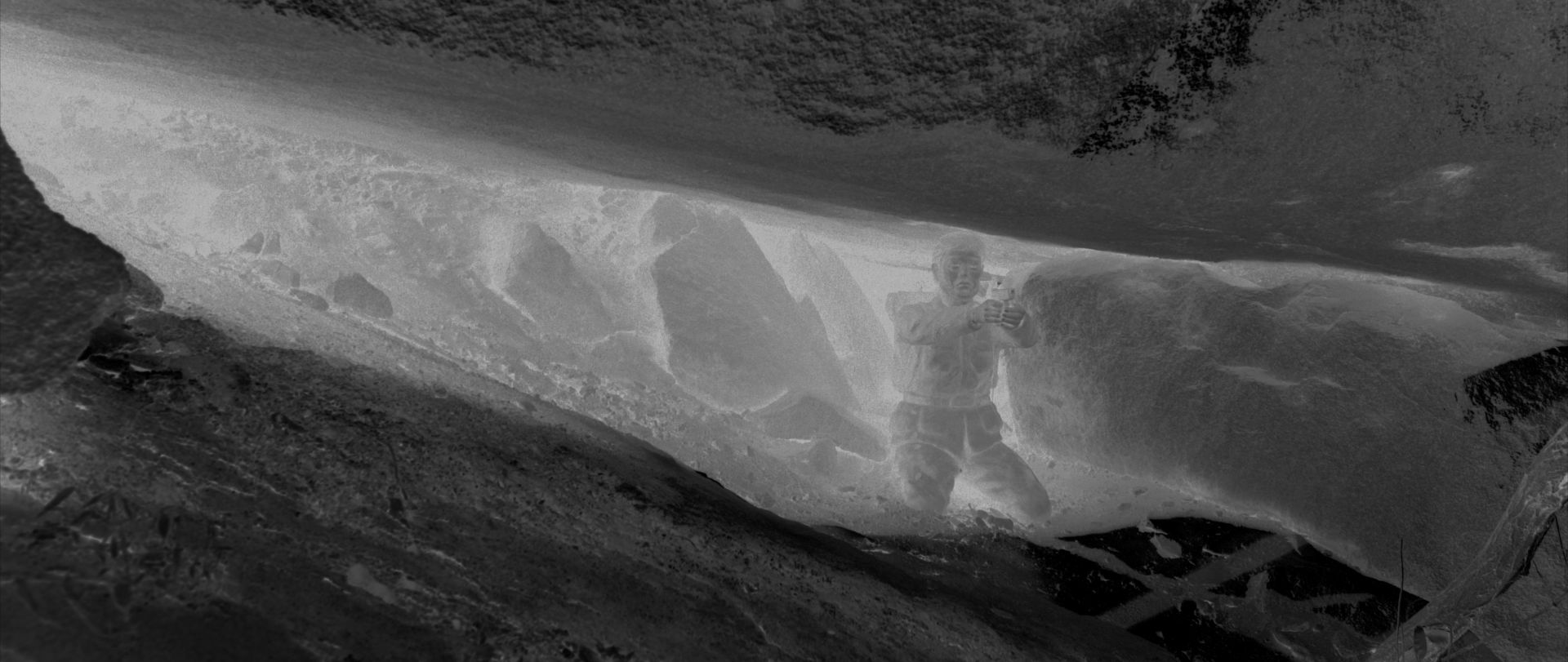 A black and white negative image features a human-like figure situated among angular and rocky forms.