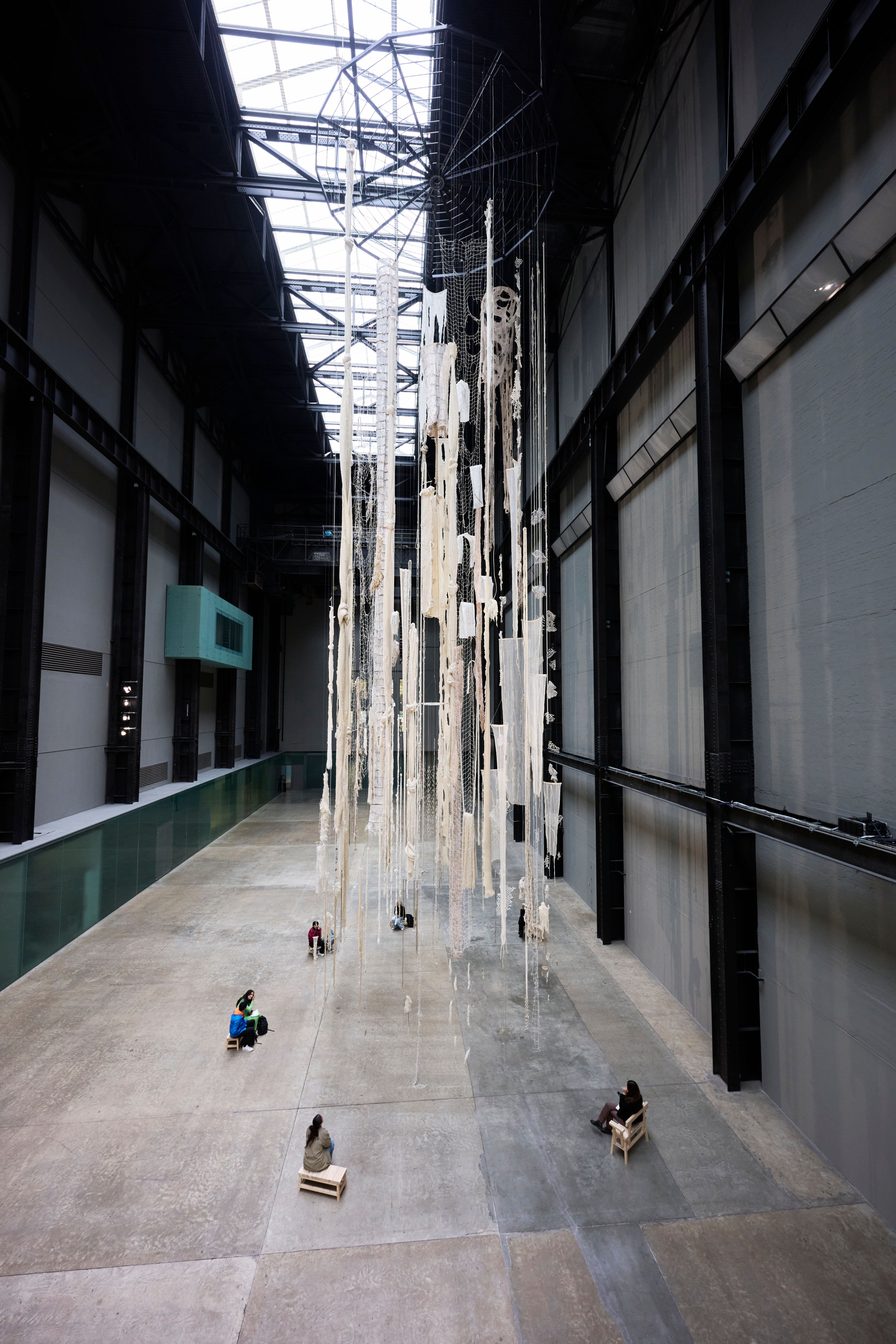People are sitting on wooden benches arranged in a circular formation beneath an art installation in a large gallery hall....