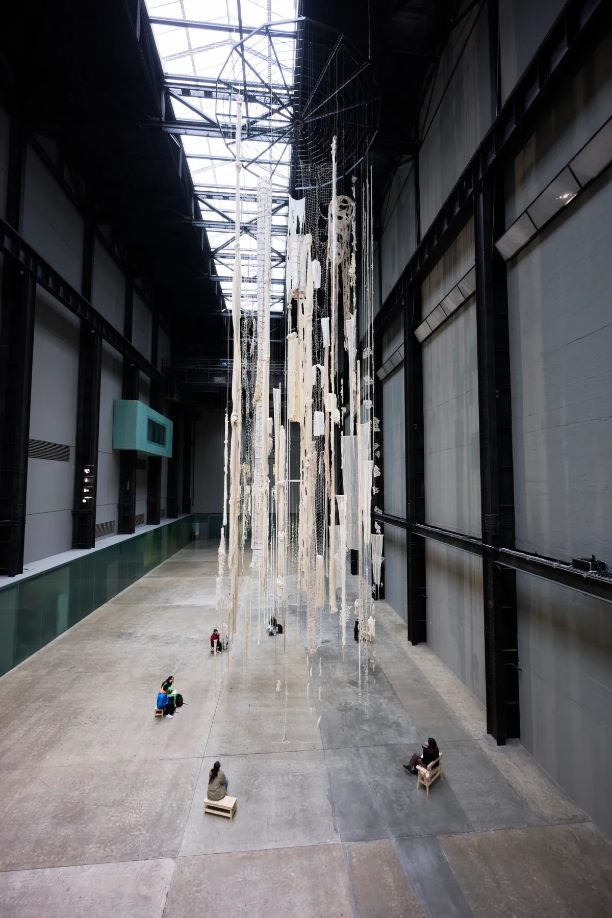 People are sitting on wooden benches arranged in a circular formation beneath an art installation in a large gallery hall. The installation consists of white, ripped curtains, fishnets, and rugs hanging from the ceiling.