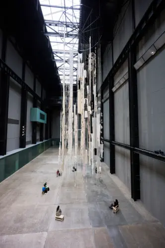 People are sitting on wooden benches arranged in a circular formation beneath an art installation in a large gallery hall....