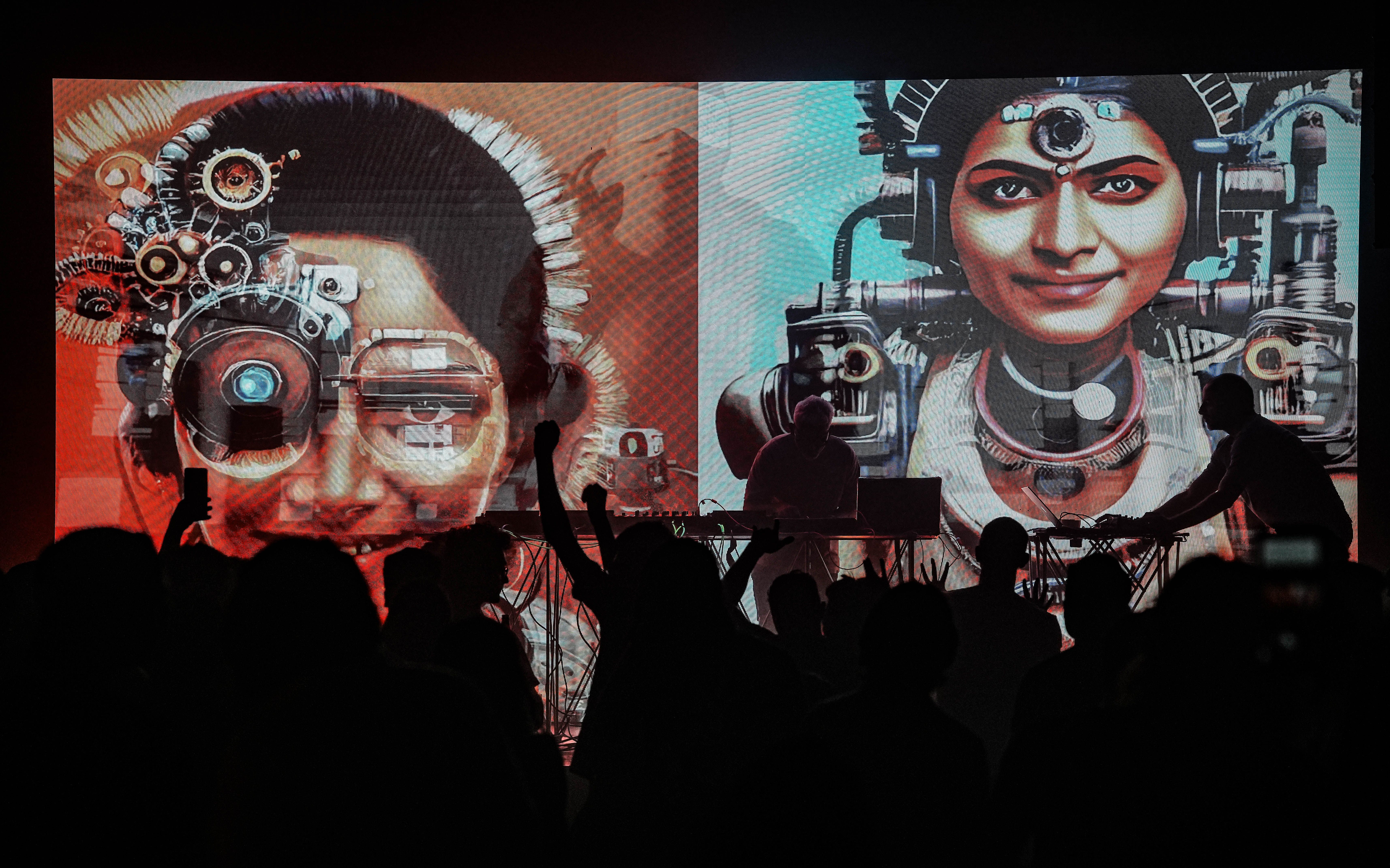 A live electronic music performance with vibrant digital art projections of cybernetic faces on a screen in the background.
