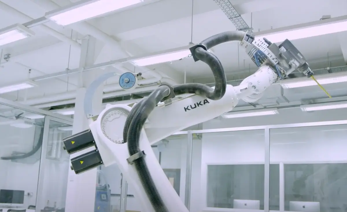 A robotic arm featuring a drill and the text "Kuka" operates in a brightly lit lab.
