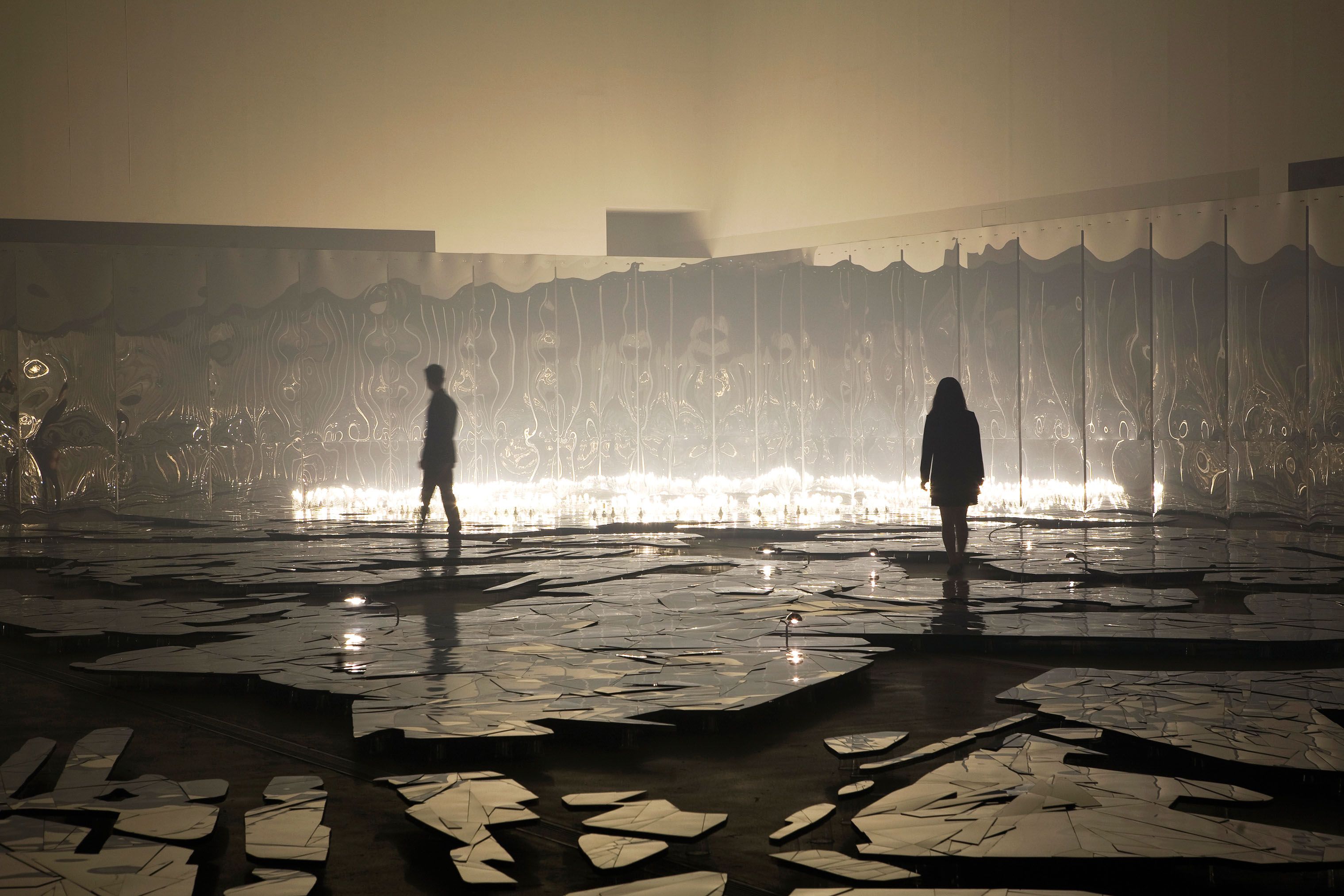 Curvy mirror walls reflect the dark showroom floor, where people are walking on a surface made to look like broken glass, ...