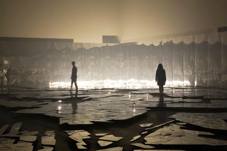Curvy mirror walls reflect the dark showroom floor, where people are walking on a surface made to look like broken glass, ...